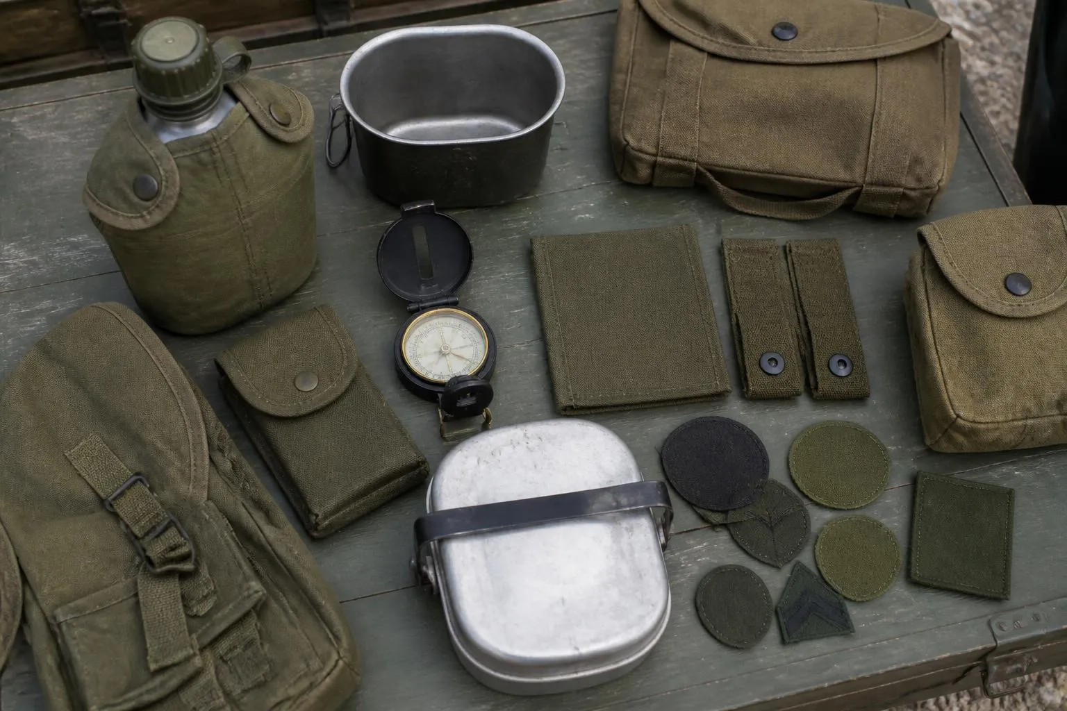 du matériel de surplus militaire étalé sur une table verte : gourde, quart en aluminium, boussole, sacoches en toile kaki et insignes